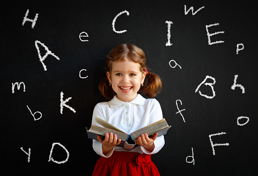 Happy schoolgirl preschool girl with book near school board blackboard