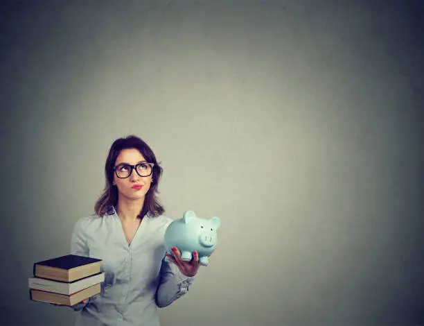 Student loan concept. Young woman with pile of books and piggy bank full of debt rethinking future career path