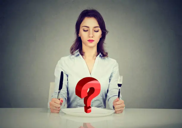 Photo of Young woman sitting at table with fork and knife looking at plate with red  question mark isolated on gray background