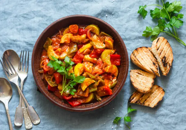 Photo of Healthy vegetarian lunch - stewed garden vegetables. Vegetable ratatouille and grilled bread. On a blue background, top view