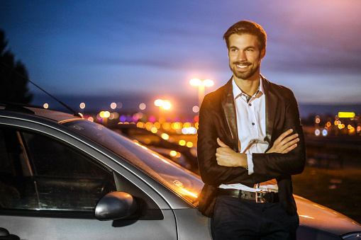 Modern businessman standing next to a limousine car at night. About 25 years old, Caucasian male.