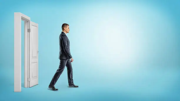 A businessman on blue background walking through an open white doorframe. New business ventures. Entering new market. Career growth.