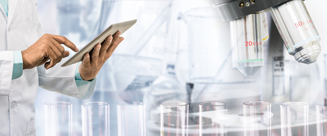 Science research and technology concept - Scientist holding tablet computer with scientific instrument, microscope and chemical test tube in lab background.