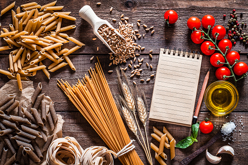 Top view of wholegrain spaghetti and penne with ingredients for cooking pasta shot on rustic wooden table. The composition includes a variety of wholegrain pasta, olive oil, tomatoes, salt, peppercorns, garlic and basil leaves. A blank note pad with useful copy space for text and/or logo is at the right. Predominant color is brown. Low key DSRL studio photo taken with Canon EOS 5D Mk II and Canon EF 100mm f/2.8L Macro IS USM