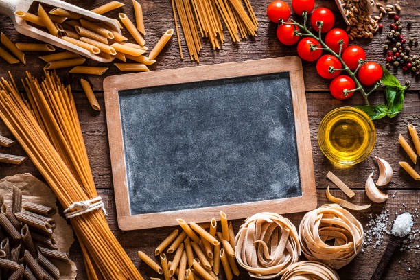 Top view of wholegrain spaghetti and penne with ingredients for cooking pasta places all around an old blank blackboard with useful copy space ready for text and/or logo shot on rustic wooden table. The composition includes a variety of wholegrain pasta, olive oil, tomatoes, salt, peppercorns, garlic and basil leaves. Predominant color is brown. Low key DSRL studio photo taken with Canon EOS 5D Mk II and Canon EF 100mm f/2.8L Macro IS USM