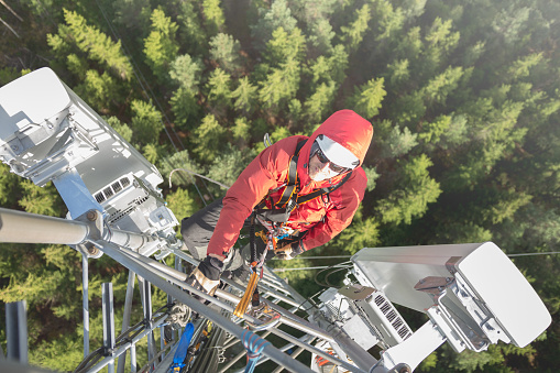 Telecommunication manual high worker engineer installing new 3g 4g LTE antenna on tall mobile base station (communication tower) in the middle of european forest, high angle of view. Working at height. Telecommunication masts and towers are typically tall structures designed to support antennas for telecommunications and broadcasting. Drone point of view.