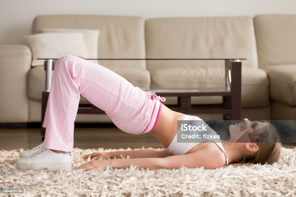 Woman in sportswear doing hip bridge exercise Joyful attractive young woman lying on floor in living room and doing hip bridge exercise. Happy smiling lady in sportswear lifting pelvis on carpet, enjoying physical activity and fitness at home Pelvis Stock Photo