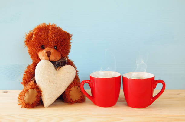 Valentines day concept. Cute teddy bear sitting and holding a heart next to couple of coffee cups, on wooden table