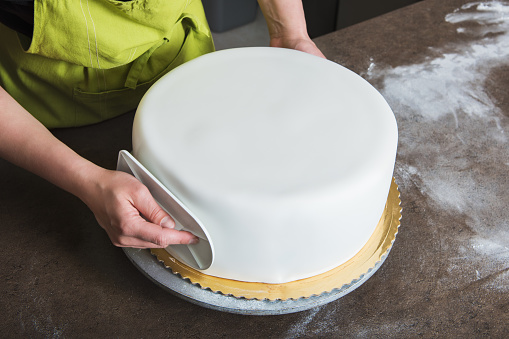 Unrecognisable woman in bakery decorating wedding cake with white fondant