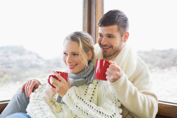 istock Couple in winter wear drinking coffee against window 836340320