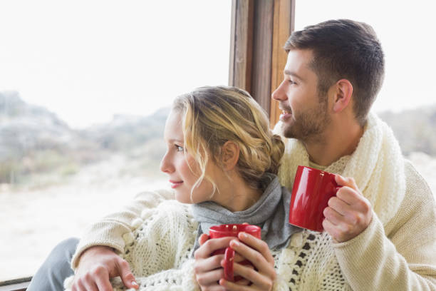 istock Couple in winter wear with cups looking out through window 836340348