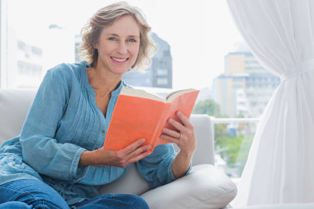 alegre mulher loira, sentado em seu sofá segurando um livro - women book mature adult reading - fotografias e filmes do acervo