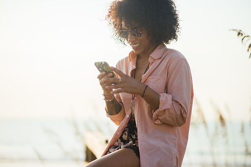 Millennial black woman texting on smartphone at beach.