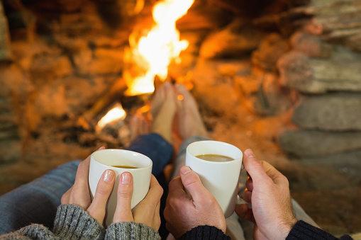 Close up of hands holding coffee cups in front of lit fireplace
