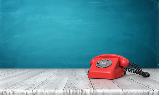 3d rendering of a bright red dial phone standing on a wooden desk and a blue wall background. Vintage appliances. Communication. Grandma phone.