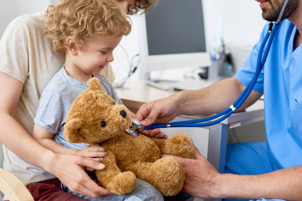 Mother and Child in Pediatric Office Portrait of adorable curly child  sitting on mothers lap in doctors office holding teddy bear toy, with pediatrician listening to heartbeat using stethoscope pediatrician stock pictures, royalty-free photos & images