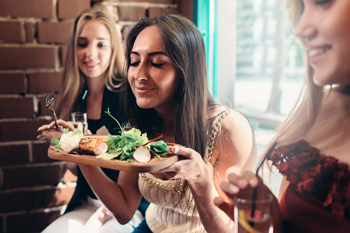 Group of girls having lunch in fashionable restaurant. Smiling young woman enjoying the smell of delicious salad served on wooden plate.