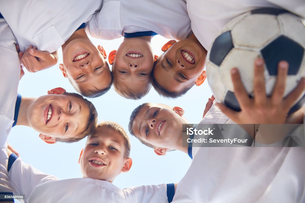 Junior Football Team Huddling Low angle portrait of junior football team standing in circle, huddling and smiling  at camera against blue sky Soccer Stock Photo