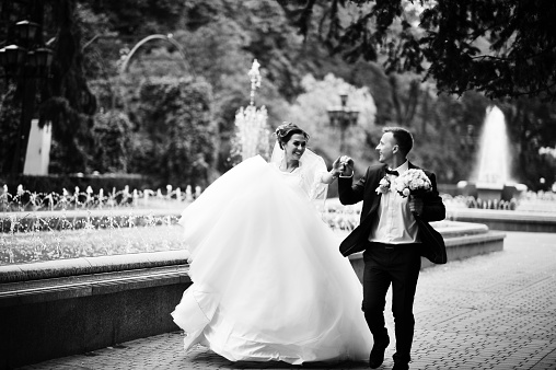 Wedding couple walking and smiling on pavement with a fountain in the background. Black and white photo.