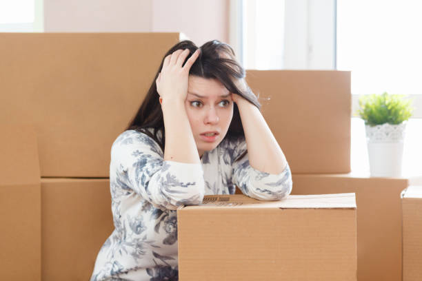 Worried and frustrated young woman sitting on packaged boxes at house for sale stock photo