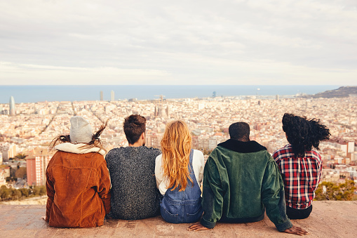 Rear view of friends sitting on building terrace. Males and females are looking at cityscape and sea. They are wearing casuals.