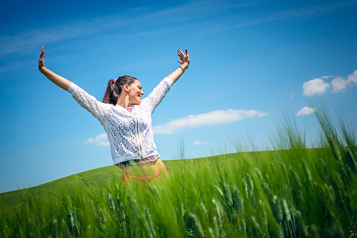 Pretty young woman with arms raised, enjoying life in young wheat field. Tuscany, Italy, Europe
