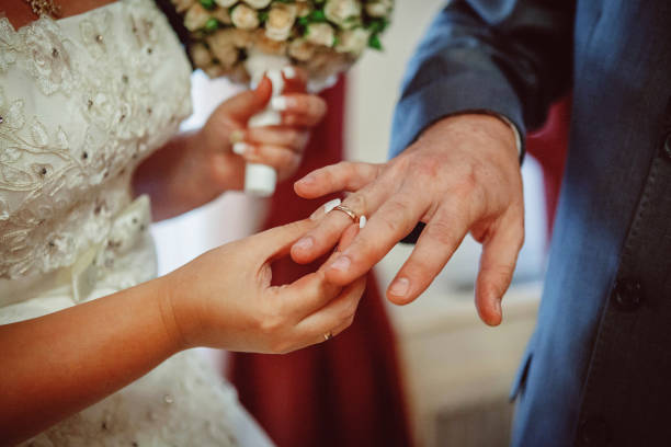 A close up shot of an exchange of rings at a wedding.  A caucasian bride dressed in a white wedding dress is placing a ring on the groom dressed in a black tuxedo.  They are holding hands as they exchange rings.  The pastor is officiating the wedding behind them and is holding  a bible.