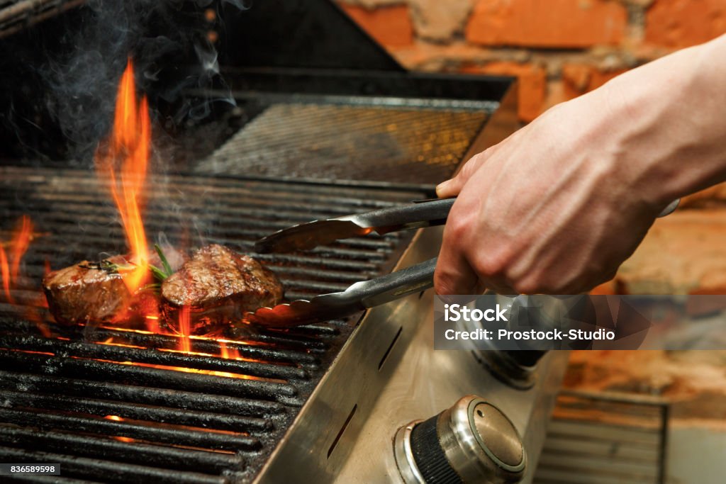 Man cooking meat steaks on professional grill outdoors Man cooking bbq meat steaks on professional grill outdoors. Male hand with tongs flipping beefsteaks on open fire Grilled Stock Photo