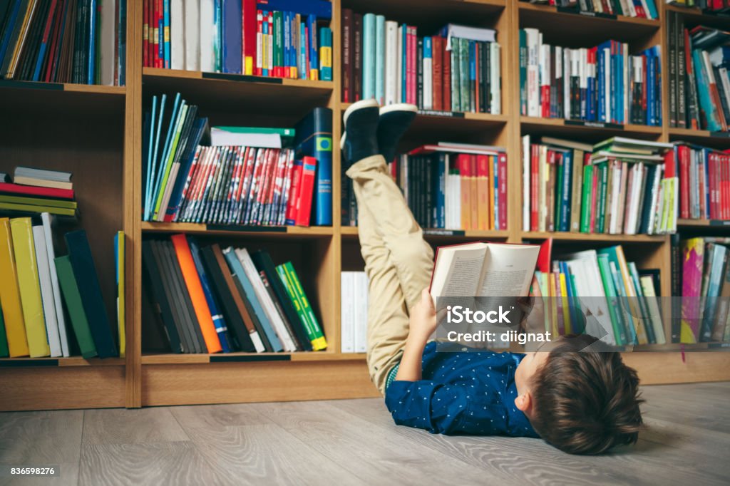 Boy in library Boy laying on the floor with the feet up, reading a book against multi colored bookshelf in library. Education, Knowledge, Bookstore, Lecture. Pupil holds a book in his hands. Child Stock Photo