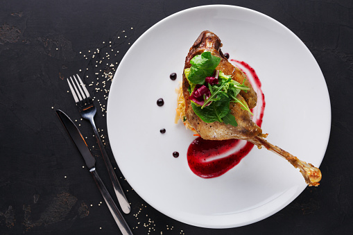 Exclusive restaurant meals. Duck confit with braised cabbage, baked apple and cranberry sauce served on snow white plate with cutlery on black table background, copy space, top view