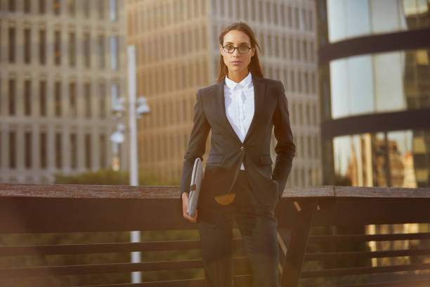 Portrait of confident businesswoman holding laptop on bridge. Young executive is standing with hand in pocket against railing. Female professional is wearing suit in city.