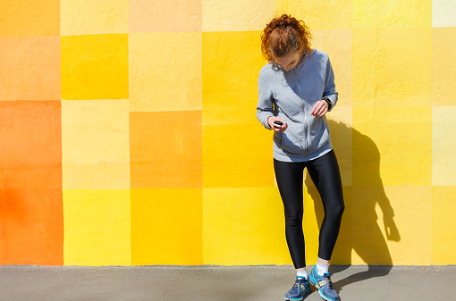 Pensive redhead woman standing at bright graffiti wall, relaxing from jog training with smartphone, copy space