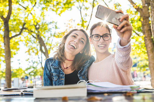 Girls studying together at park and taking a funny selfie. Happy best friends with books having fun while studying. Friendship and lifestyle concepts.