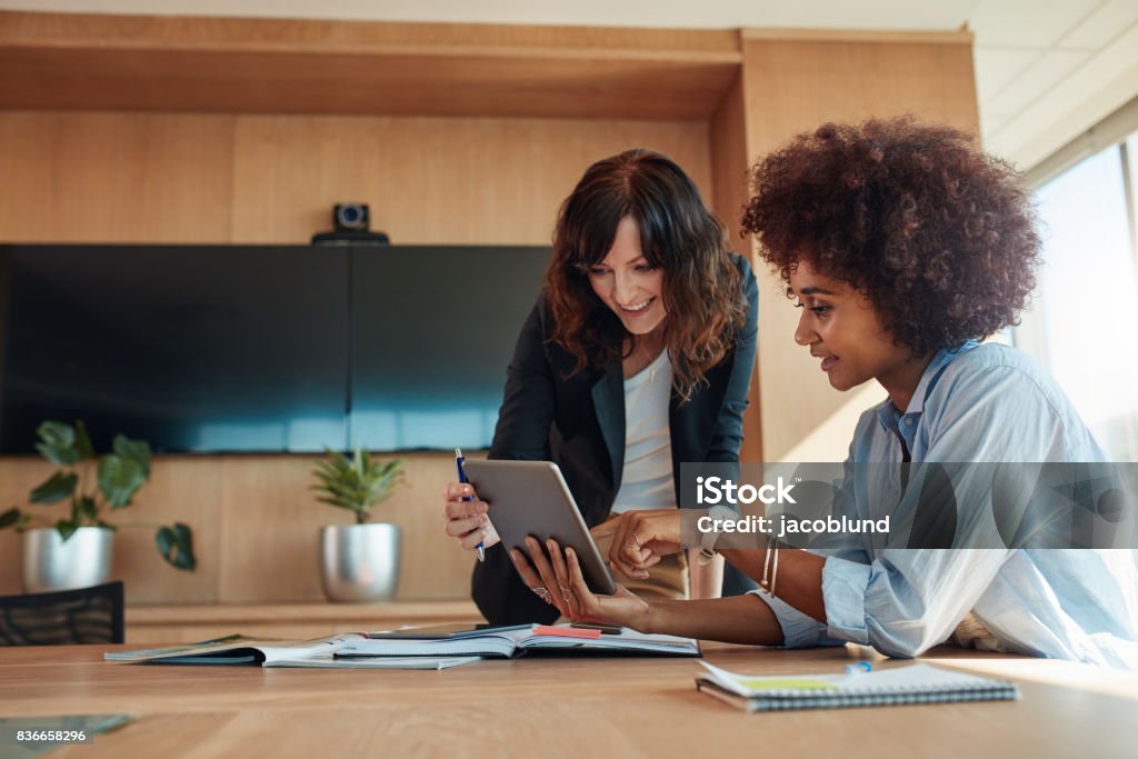 Professional employees discussing project on tablet pc African businesswoman showing something on digital tablet to her female colleague while sitting at her desk. Professional employees discussing ideas of project on tablet pc. Business Meeting Stock Photo