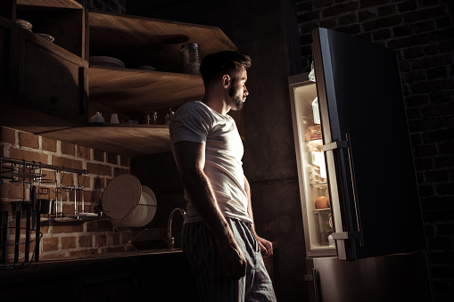 side view of bearded young man in pajamas looking at open refrigerator at night