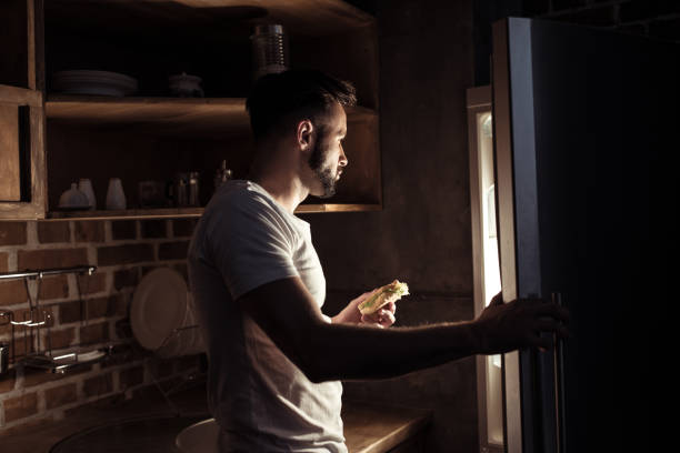 young man in pajamas eating and looking at refrigerator at night