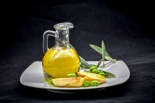 a compisition made by a jar full of oil, an olive branch, some olives and an italian frisella on a black background