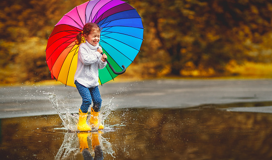 Happy funny ba child by girl with a multicolored umbrella jumping on puddles in rubber boots and laughing