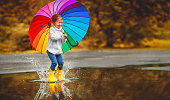 Happy funny child girl with  umbrella jumping on puddles in rubber boots