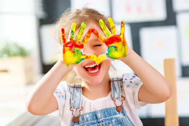 Photo of funny child girl draws laughing shows hands dirty with paint