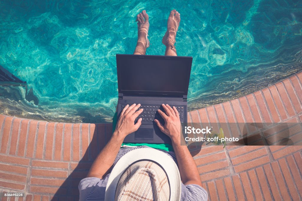 Tourist sitting at the edge of swimming pool and using blank screen laptop. Summer vacation and freelancer concepts High angle view of man sitting at the poolside and using laptop. Travel and technology concepts Swimming Pool Stock Photo