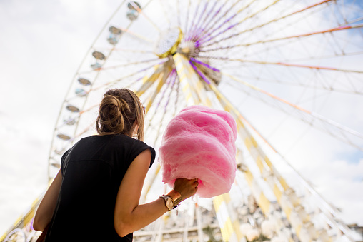 Young woman standing with pink cotton candy in front of the ferris wheel at the amusement park