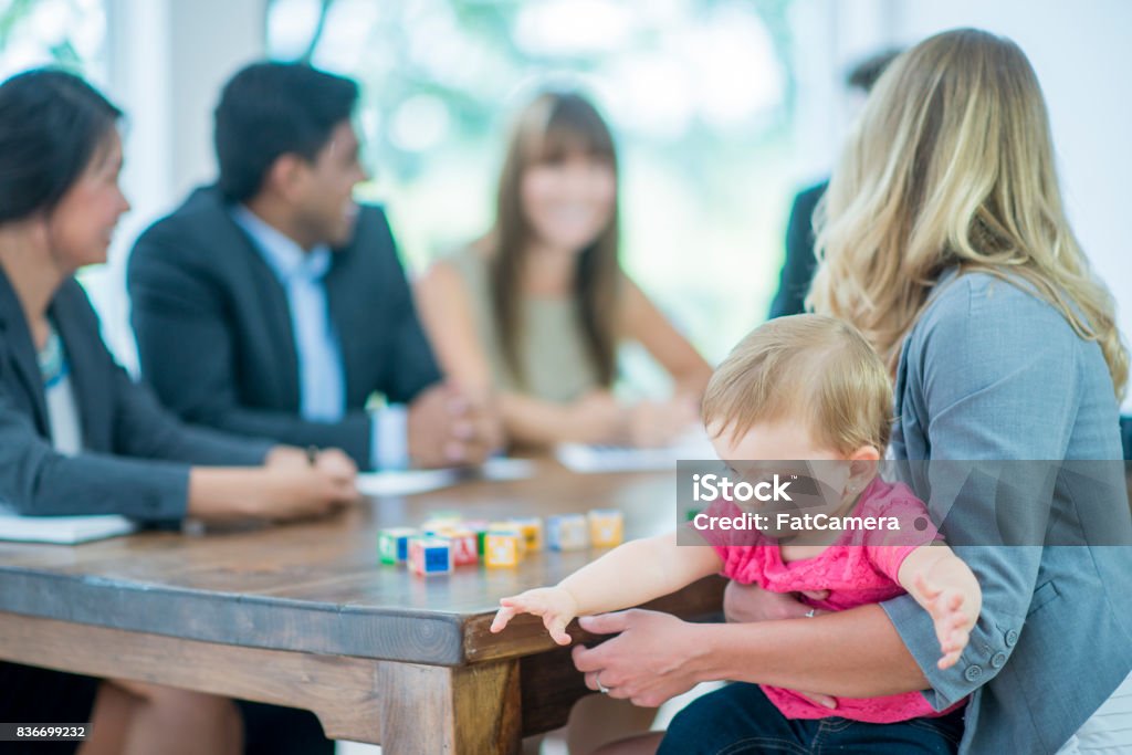 Work And Play A multi-ethnic group of adults business-people are indoors in a board room. They are wearing formal clothing. A Caucasian woman is holding her baby during a meeting. The baby is playing with blocks. Imbalance Stock Photo