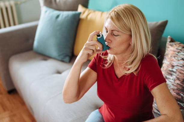 Daily Life of a Person with Asthma Senior woman in her late 50s inhaling asthmatic cure at home. Woman is living life with chronic illness everyday and overcoming challenges with it. asthma inhaler stock pictures, royalty-free photos & images