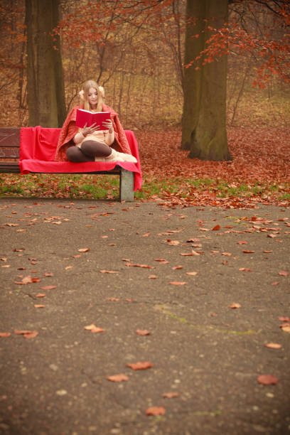 Outdoor nature season relax leisure concept. Young lady with literature. Blonde girl reading red book sitting on bench in autumnal woodland.
