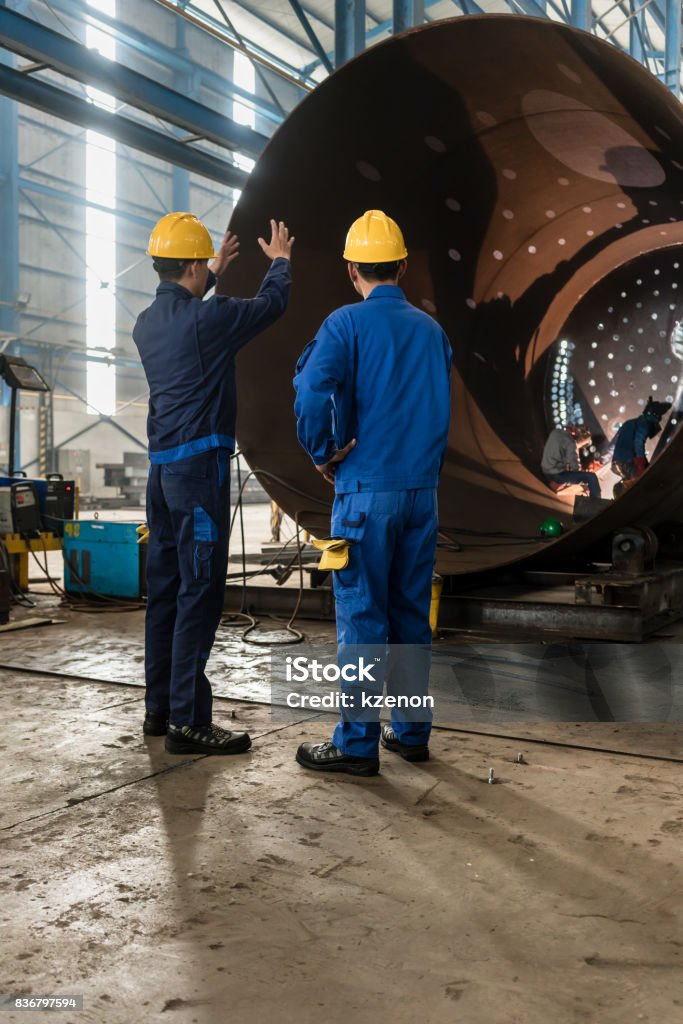 Workers supervising the manufacture of a metallic cylinder Two experienced workers supervising the manufacture of a metallic cylinder in the interior of a factory Occupation Stock Photo