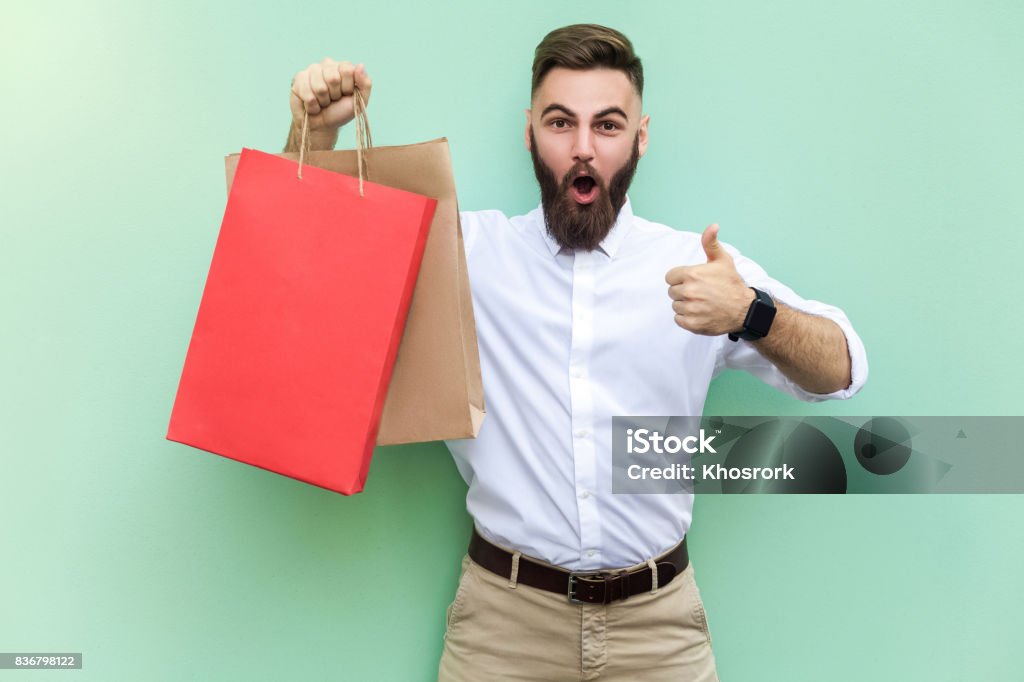 Online shopping. Young adult businessman thumbs up and looking at camera with surprised face. Online shopping. Young adult businessman thumbs up and looking at camera with surprised face. Indoor, studio shot on light green background Men Stock Photo