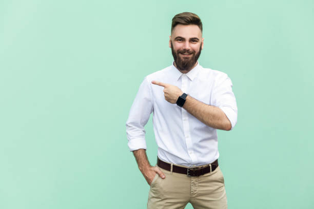 espace copie pointage par homme d’affaires. beau jeune homme adulte avec barbe en chemise blanche, regardant la caméra et en pointant loin en se tenant debout, isolé sur fond blanc - joueur de champ gauche photos et images de collection