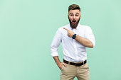 Handsome young adult man with beard in shoked. Pointing away while standing isolated on light green background