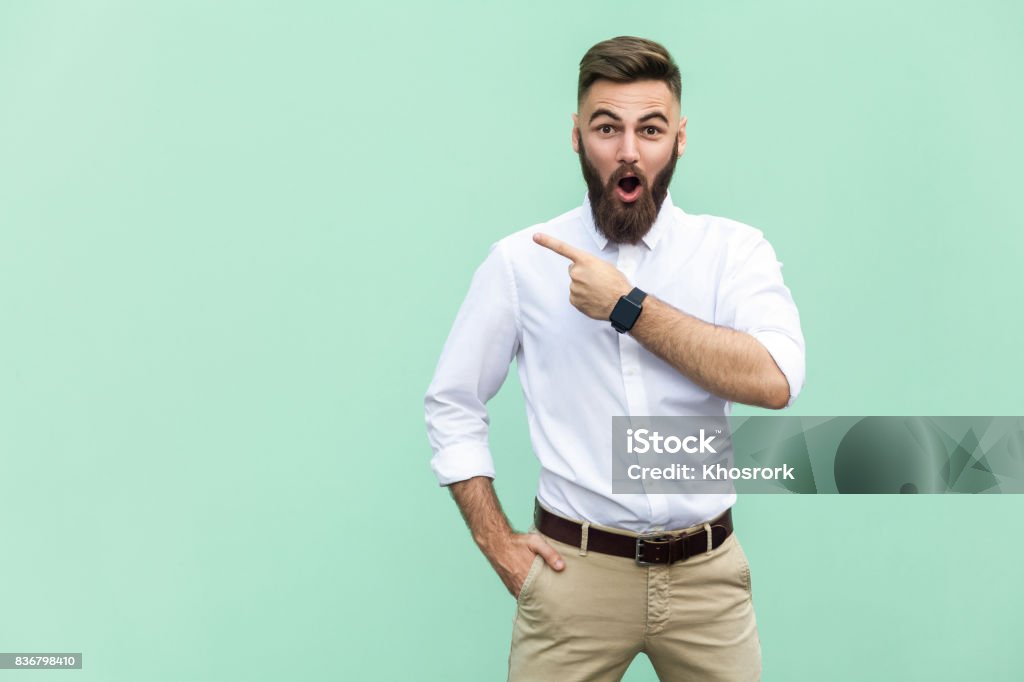Handsome young adult man with beard in shoked. Pointing away while standing isolated on light green background Men Stock Photo